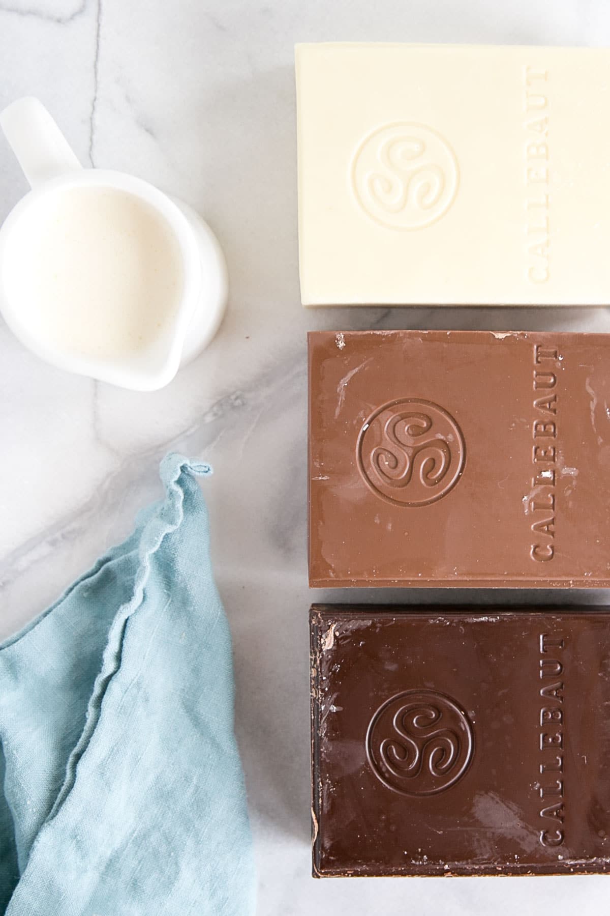 How to make Chocolate Ganache! A simple, two-ingredient recipe for a delicious frosting, filling, drip, or glaze. | livforcake.com White, milk, and dark chocolate bars beside a small pitcher of cream.