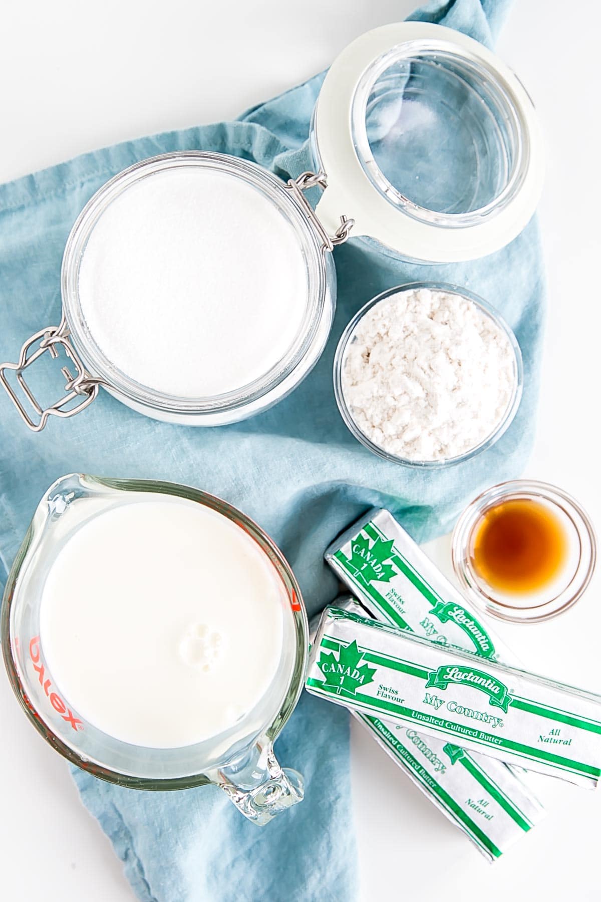 Ermine Frosting (also known as flour buttercream or heritage frosting) is an easy and delicious buttercream made with flour, sugar, milk, and butter. | livforcake.com Ingredients shown against a blue tea towel -- milk, sugar, flour, butter, and vanilla.
